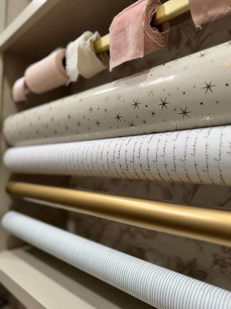 Gold rods holding pastel wrapping paper and ribbon rolls in custom gift wrapping closet by JKE Home Reset.